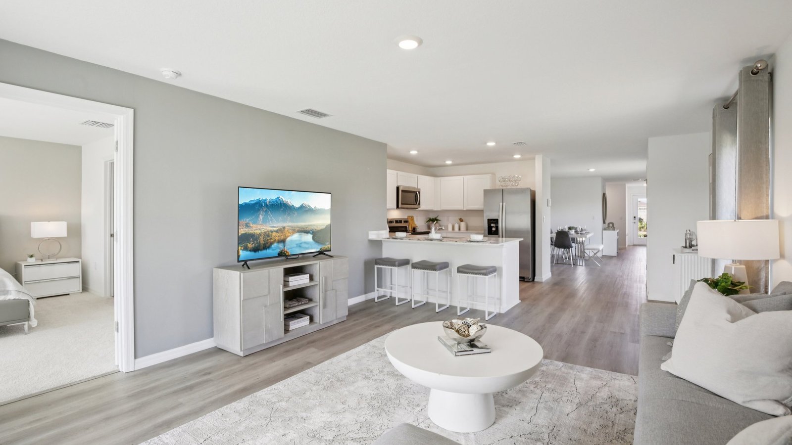 Modern open-plan living room with light gray walls, a sofa, round white coffee table, TV, kitchen with white cabinets and bar stools, and a doorway leading to a bedroom with a bed and nightstand.