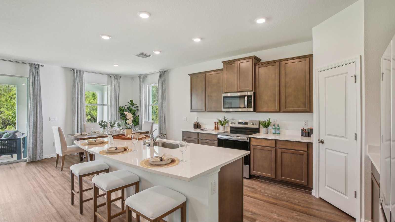 Modern kitchen with wood cabinets, stainless steel appliances, and white countertops. An island with stools faces a dining area with a set table. Large windows bring in natural light and greenery can be seen outside.
