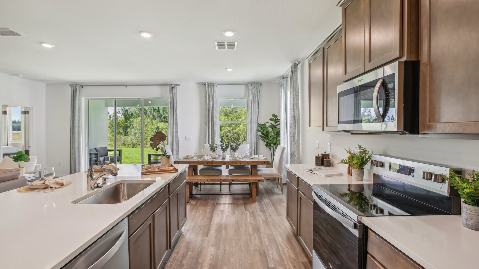 Modern kitchen with white countertops, stainless steel appliances, and wooden cabinets. In the background, a dining table is set next to large windows and sliding glass doors overlooking a green outdoor area.