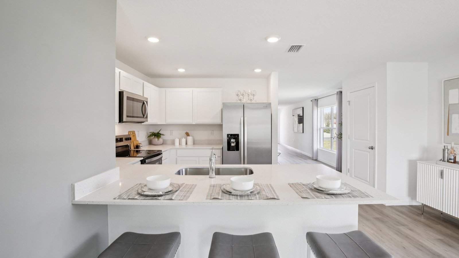 Modern kitchen with white cabinets, stainless steel appliances, a large refrigerator, and a kitchen island with three stools and place settings. The space is bright with natural light from nearby windows.