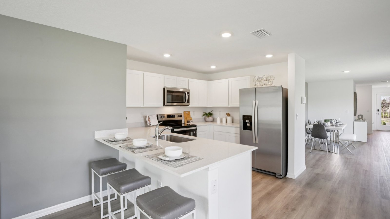 Modern kitchen with white cabinets, stainless steel appliances, a large refrigerator, and a breakfast bar with three gray cushioned stools. The space opens to a dining area with wood flooring and neutral decor.