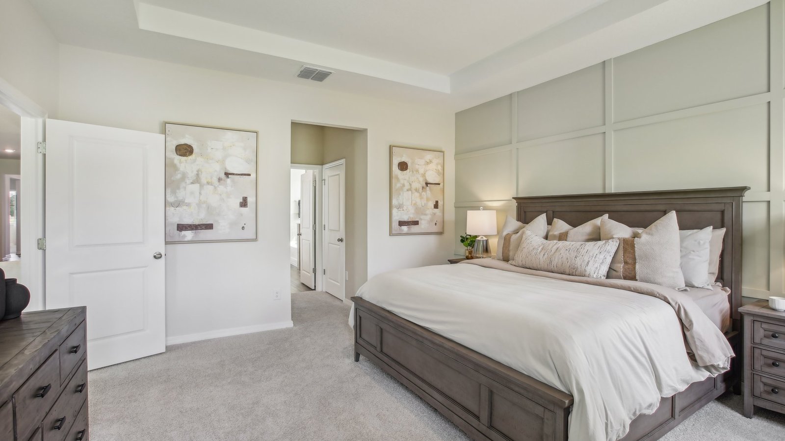A modern bedroom with a large wooden bed, neutral bedding, two abstract paintings on the wall, a nightstand with a lamp, dresser, and an open door leading to a bathroom. Walls and carpet are light-toned.