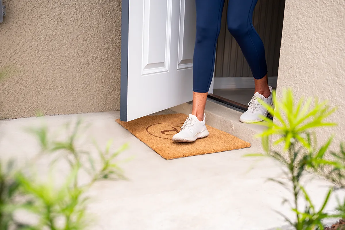 A person wearing white sneakers and navy leggings steps out of a door onto a tan doormat with a smiley face, surrounded by light green plants.