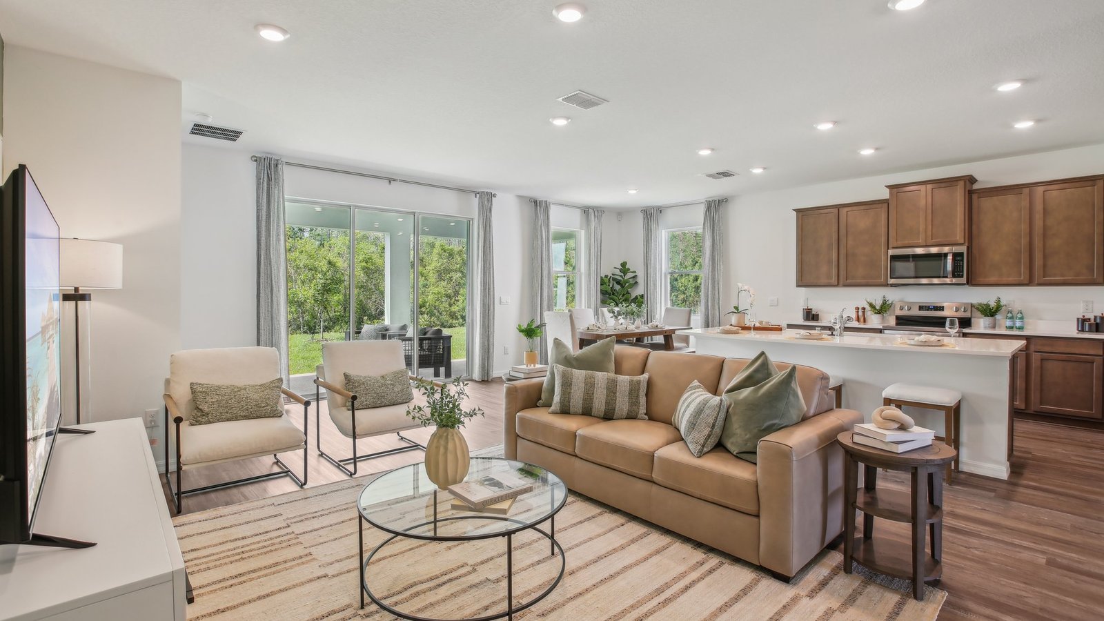 A modern open-concept living room and kitchen with neutral tones, featuring a tan sofa, two armchairs, a glass coffee table, wood cabinets, stainless steel appliances, and large windows with views of greenery.