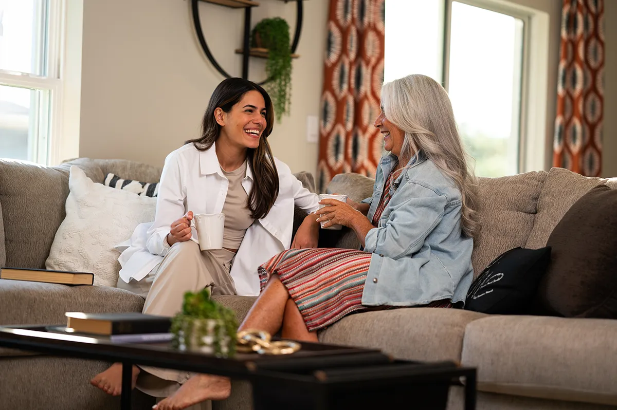 Two women sit on a couch in a cozy living room, smiling and holding mugs as they chat. One wears a light coat and pants, the other wears a striped dress and has long gray hair.