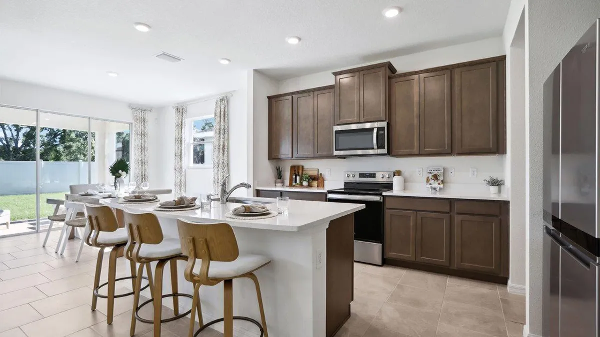 Modern kitchen with brown cabinets, stainless steel appliances, and a white island with four wooden bar stools. Large windows and sliding glass doors provide natural light, and a dining table is set nearby.