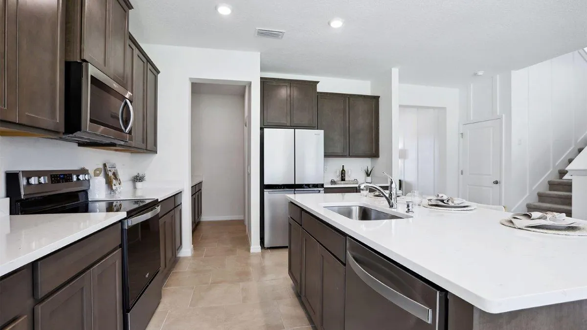 Modern kitchen with dark wood cabinets, stainless steel appliances, a center island with a sink, and white countertops. Neutral tile flooring and bright lighting create a clean, inviting space. Stairs are visible in the background.