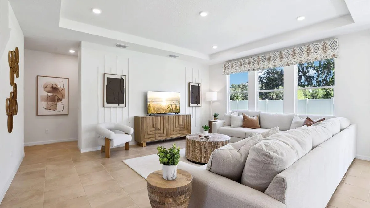 Modern living room with a large beige sectional sofa, wooden coffee tables, a TV on a console, wall art, and large windows letting in natural light. Decor includes plants, a white armchair, and neutral accents.