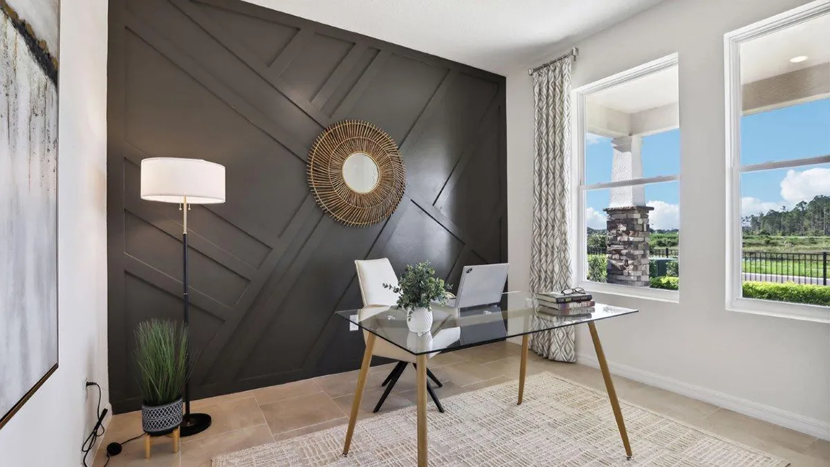 Modern home office with a glass desk, two white chairs, a laptop, books, and a potted plant. Dark accent wall with a round woven mirror, floor lamp, and large windows letting in natural light.