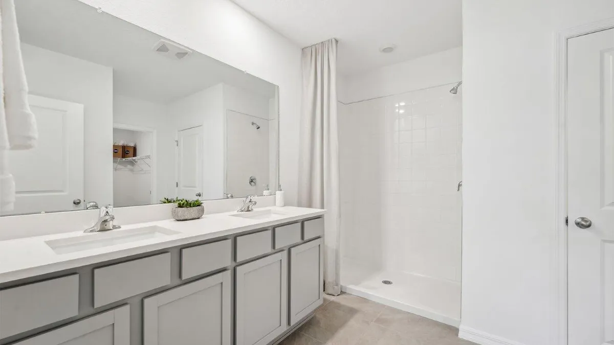Modern bathroom with a double-sink vanity, large mirror, potted plant, and a walk-in shower with a white curtain. The room features white walls and light-colored cabinetry.