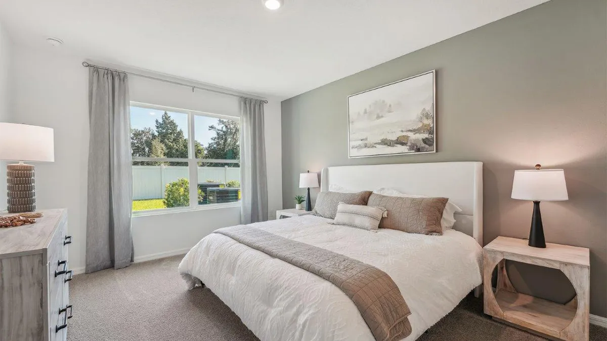 A bright bedroom with a large window, a bed with white and beige bedding, two bedside tables with lamps, a dresser, and a framed landscape painting on a gray accent wall.