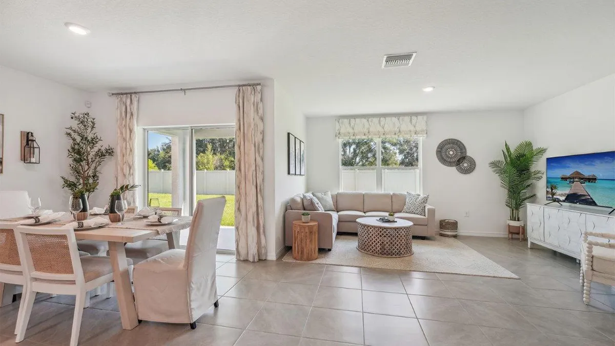 A bright, modern living and dining area with neutral decor, tiled floor, a dining table set for four, beige sofa, round coffee table, indoor plants, and a TV displaying a beach scene. Large windows let in natural light.