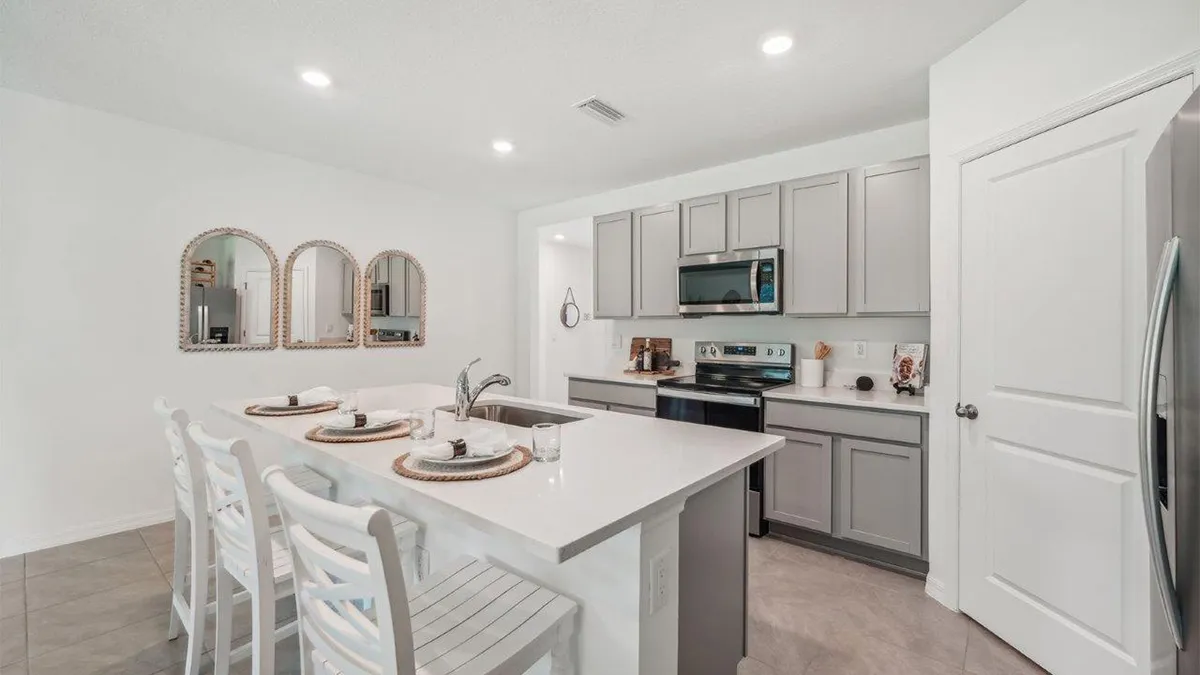 Modern kitchen with light gray cabinets, stainless steel appliances, a white island with a sink, three white chairs, place settings, and three decorative mirrors on the wall. The space is bright and uncluttered.