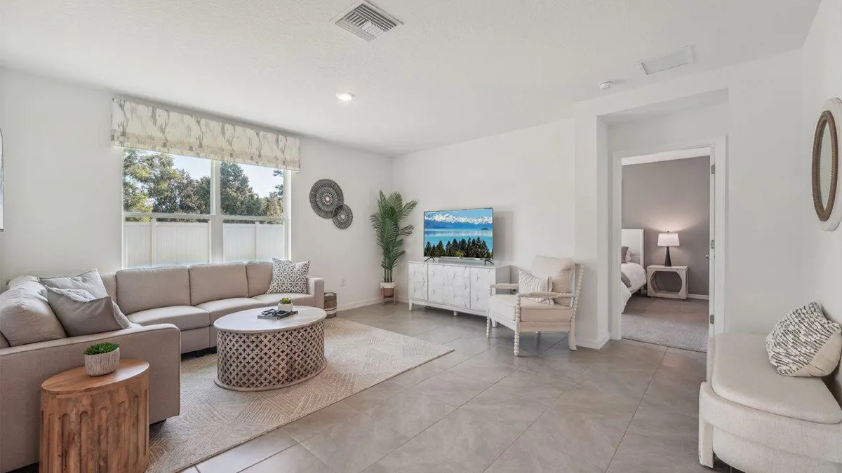 Bright, modern living room with a beige sectional sofa, round coffee table, TV on a white console, and neutral decor. Large window lets in natural light; bedroom visible through an open door.