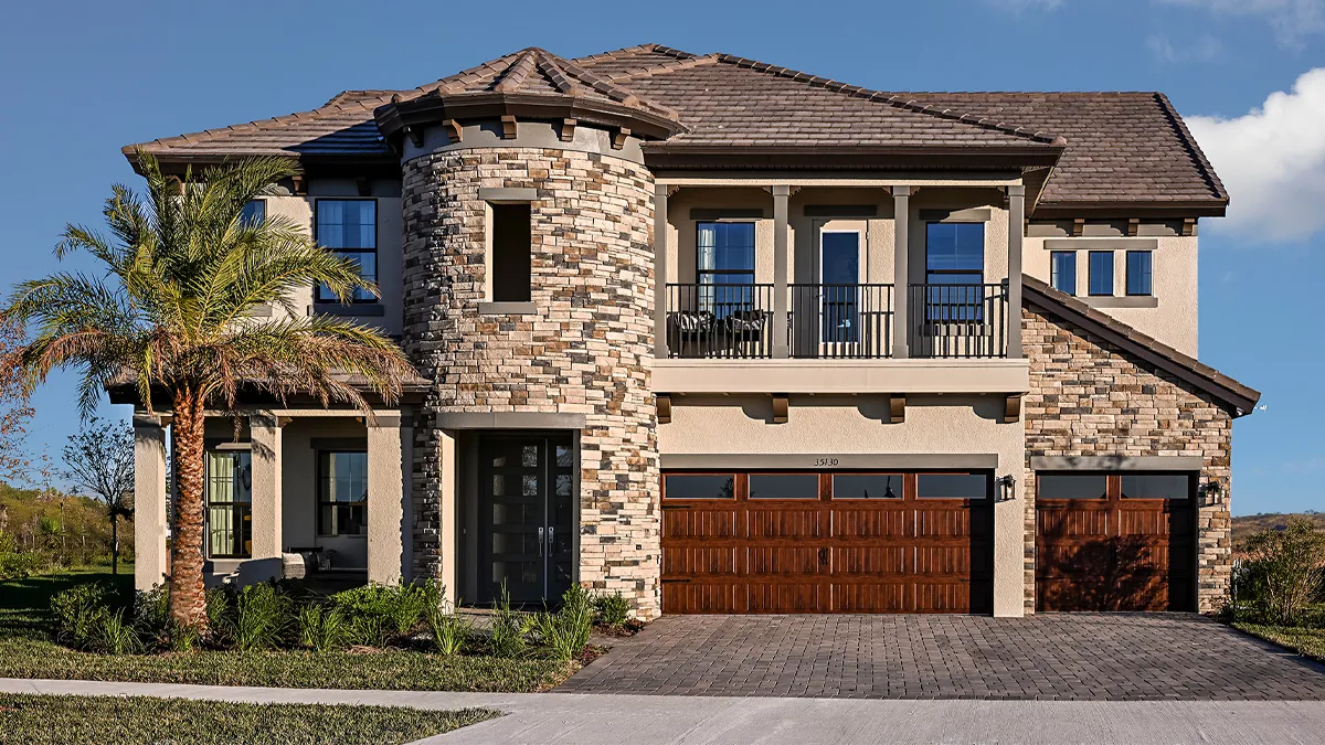 Two-story house with a stone tower feature, beige walls, dark roof tiles, wooden garage doors, front balcony with wrought-iron railing, palm tree, and landscaped lawn under a clear blue sky.