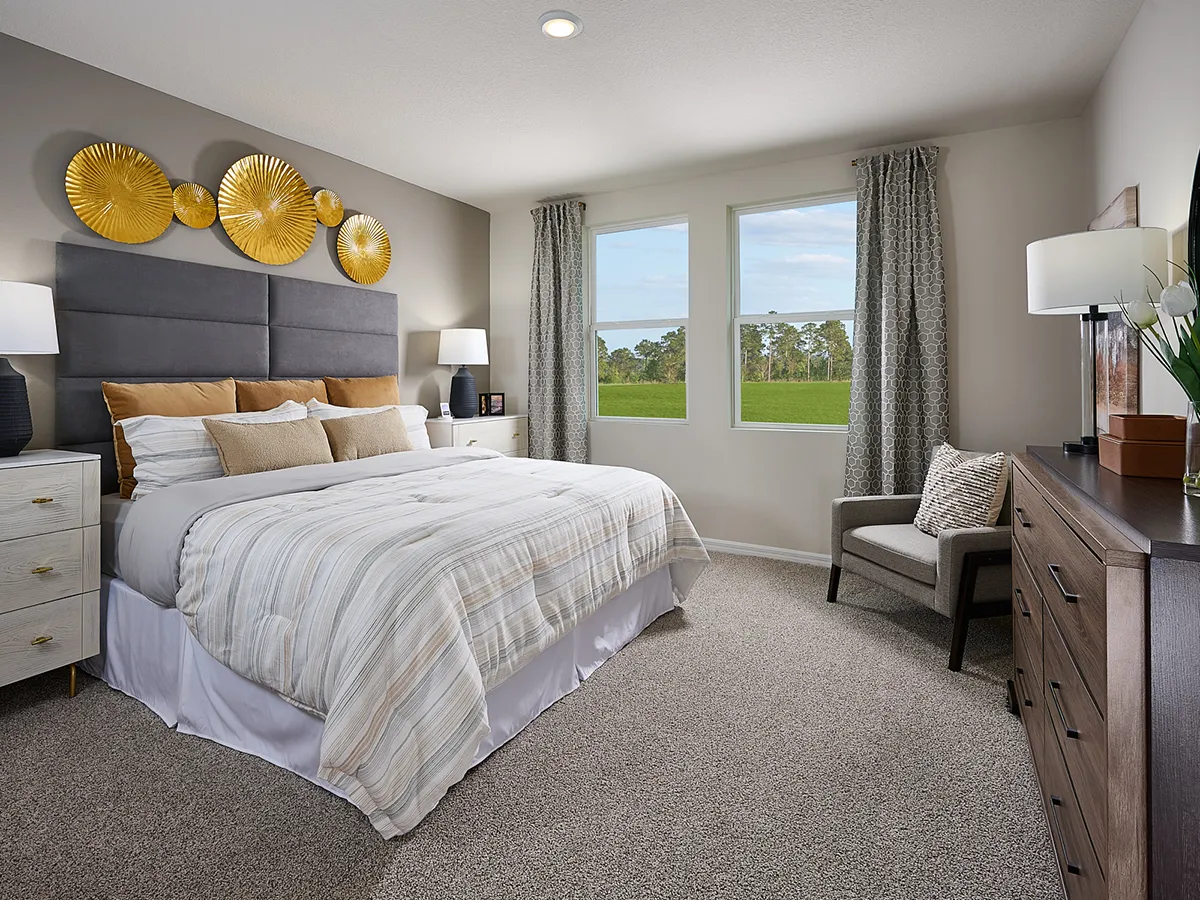 Modern bedroom with a large bed, neutral bedding, gray padded headboard, gold wall art, bedside tables with lamps, an armchair, dresser, and windows with gray curtains overlooking a green landscape.