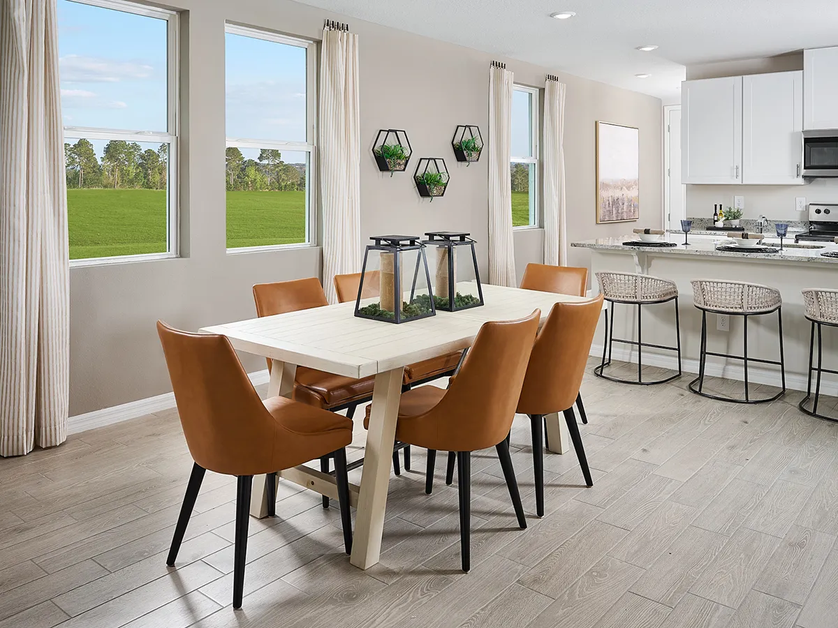 Modern dining area with a light wood table, six brown leather chairs, and decorative terrariums as centerpieces. Large windows show a green field. Nearby is a kitchen with white cabinets and bar stools.