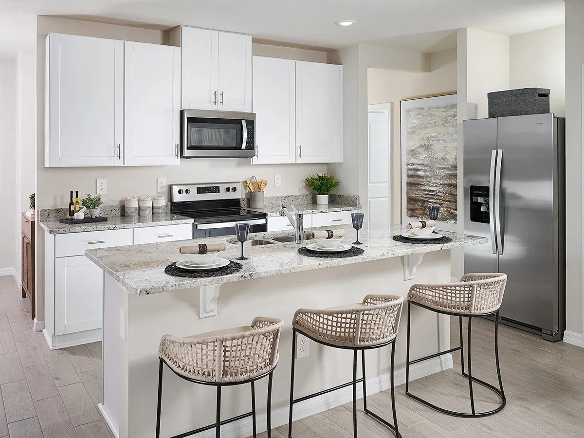 Modern kitchen with white cabinets, stainless steel appliances, a marble countertop island set with plates and glasses, and three woven barstools. Light wood flooring and decorative plants complete the space.