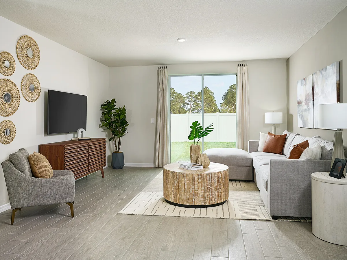 A modern living room with light wood flooring, a gray sectional sofa, a round wicker coffee table, a TV on a wooden console, wall art, and large windows with beige curtains overlooking a green yard.