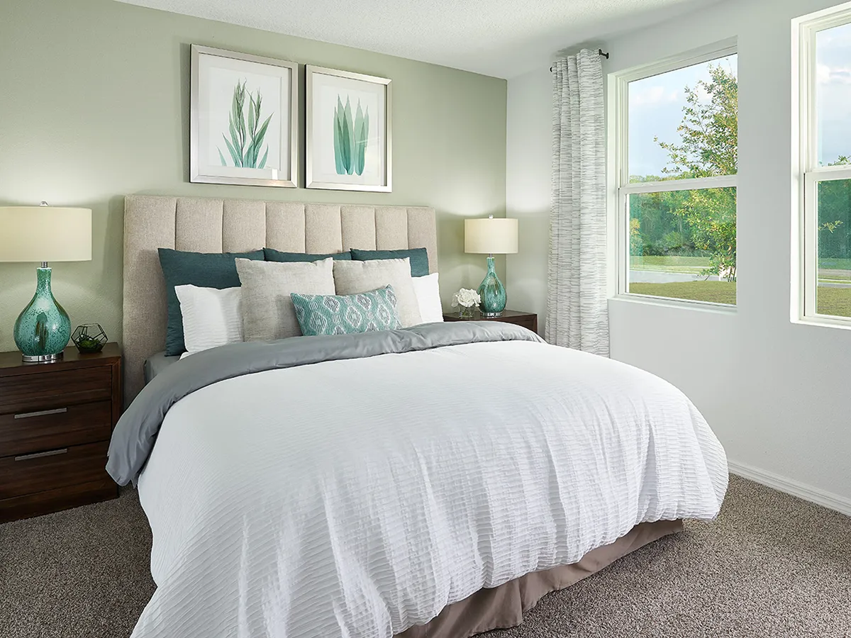 A modern bedroom with a large bed, white comforter, and multiple pillows. Two bedside tables with teal lamps and drawers flank the bed. Two plant art prints hang above the tufted headboard. Sunlight streams through two windows.