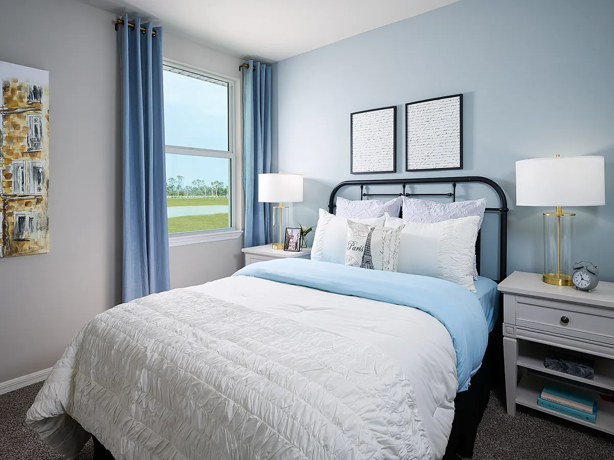 Bright bedroom with a neatly made bed featuring white and light blue bedding, two bedside tables with lamps, a window with blue curtains, and framed art on light blue walls.