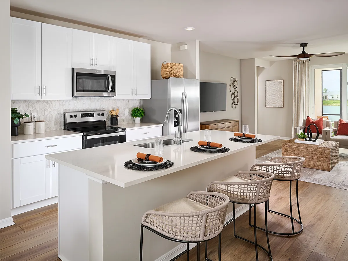 Modern kitchen with white cabinets, stainless steel appliances, a large island with a sink, and three woven barstools. Place settings with orange napkins are on the island. Living room is visible in the background.