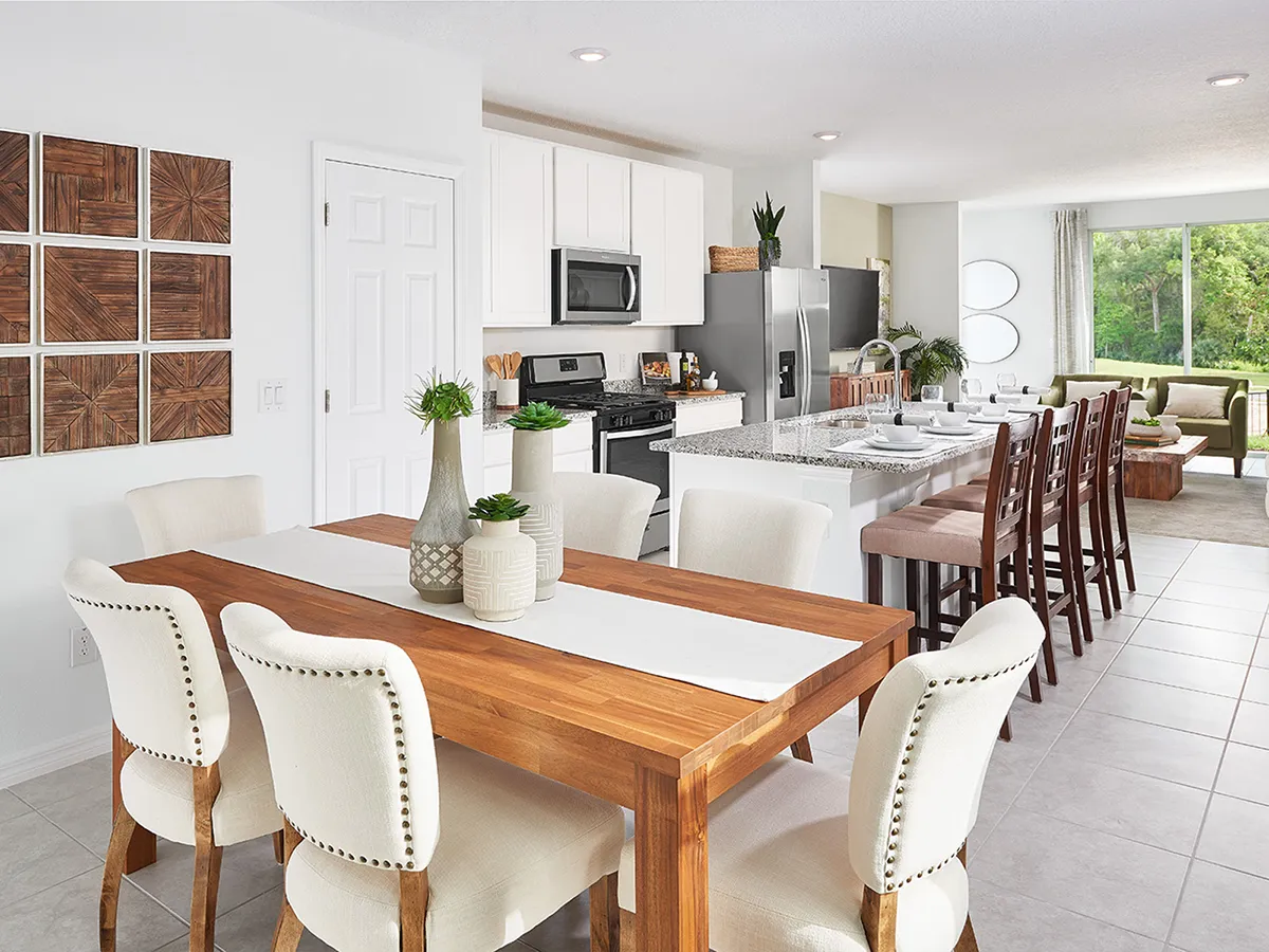 Modern open-plan kitchen and dining area with a wooden dining table, white upholstered chairs, granite kitchen island with bar stools, stainless steel appliances, and large windows letting in natural light.