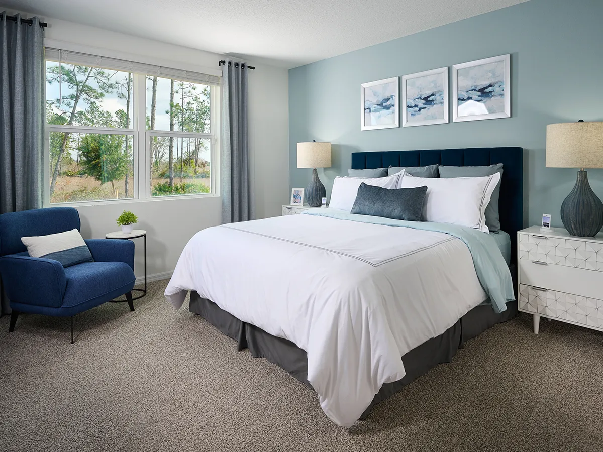 A modern bedroom with a large bed, blue headboard, white bedding, two nightstands with lamps, a blue armchair, and three abstract paintings on a light blue wall. A window with gray curtains lets in natural light.