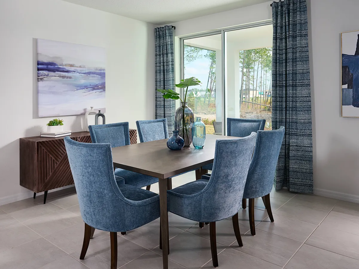 A modern dining room with a gray rectangular table, six blue upholstered chairs, blue-gray curtains, and large glass doors showing trees outside. Abstract art and a sideboard decorate the white walls.