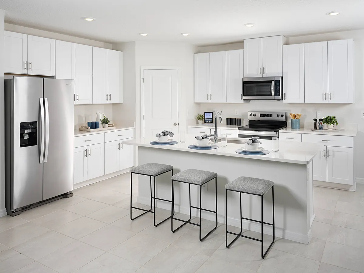 Modern kitchen with white cabinets, stainless steel appliances, a center island with three gray cushioned stools, and place settings. The room is bright with recessed lighting and minimalistic decor.