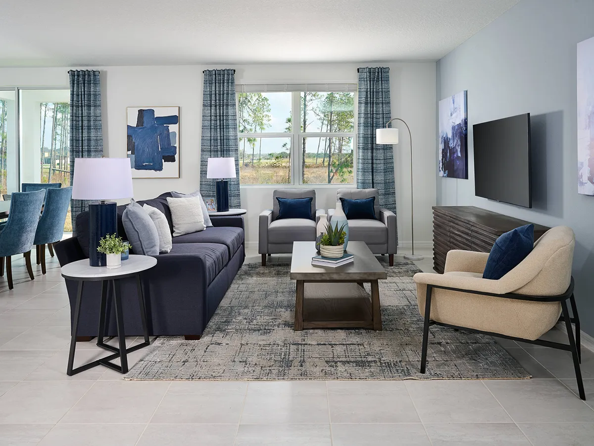 Modern living room with blue and neutral tones, featuring a dark sofa, beige armchair, two gray chairs, a coffee table with decor, floor lamp, wall art, TV, and large window with blue curtains overlooking trees.