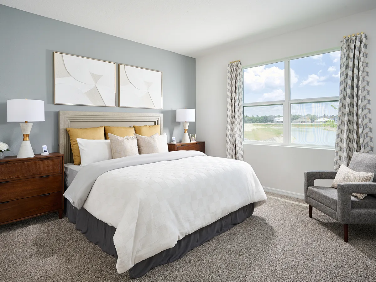 A modern bedroom with a large bed featuring white and beige bedding, two wooden nightstands with lamps, a gray accent wall, two abstract artworks, a gray armchair, and a window with patterned curtains letting in sunlight.