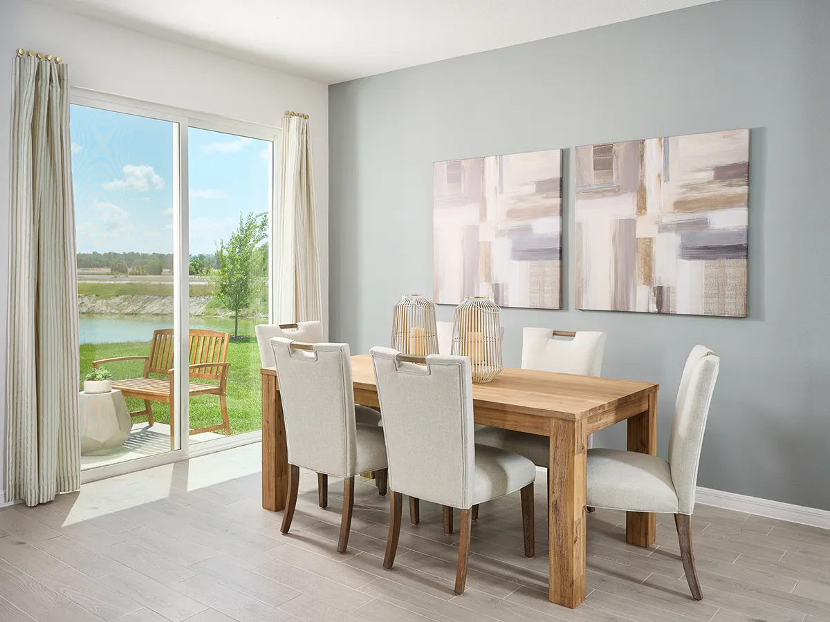 A modern dining room with a wooden table, six upholstered chairs, abstract art on a gray wall, and large glass doors opening to a patio with outdoor seating and a grassy yard by a lake.