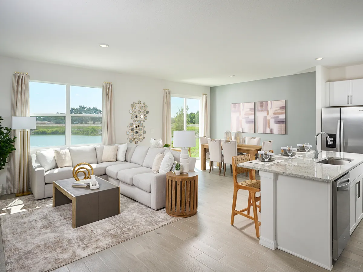 A modern, open-concept living and dining area with light-colored furniture, a large sectional sofa, a wooden coffee table, a kitchen island with stools, and large windows letting in natural light.