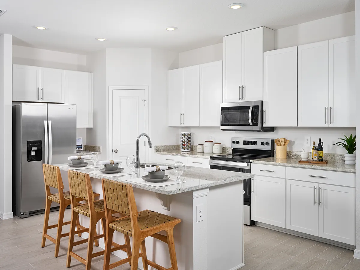 Modern kitchen with white cabinets, stainless steel appliances, and a large island with a sink. Three woven barstools are placed at the island, which is set with dishes and bowls. The space is bright and tidy.