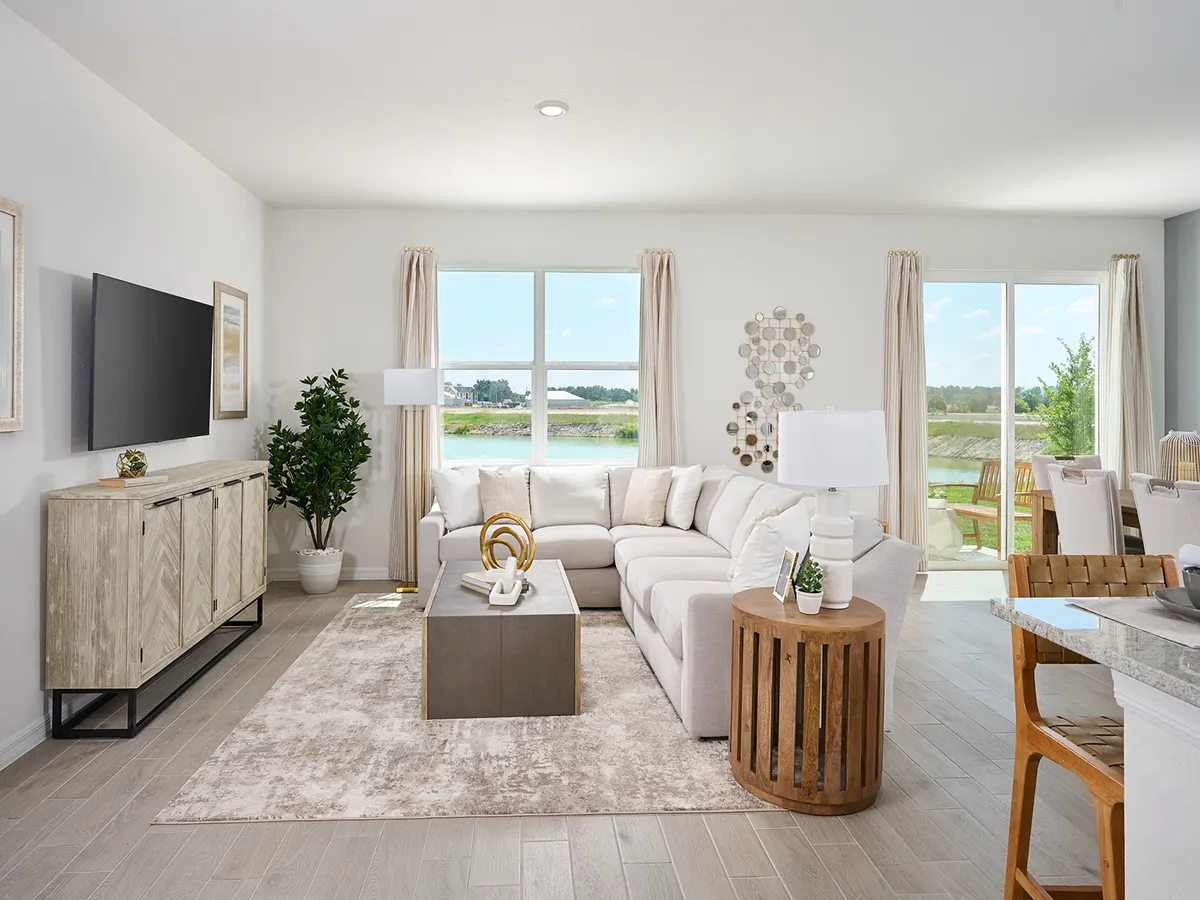 A modern, bright living room with a large beige sectional sofa, neutral rug, wooden side tables, a flat-screen TV on a wood console, green plant, and large windows showing a view of a pond and greenery outside.
