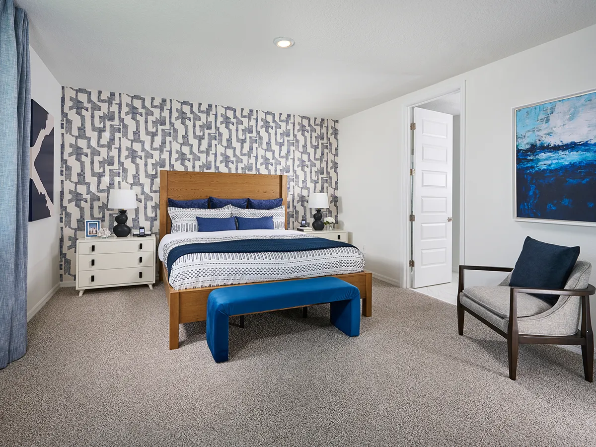 Modern bedroom with a geometric accent wall, wooden bed with blue and white bedding, two white nightstands, a blue bench at the foot of the bed, an armchair, abstract wall art, and neutral carpeting.