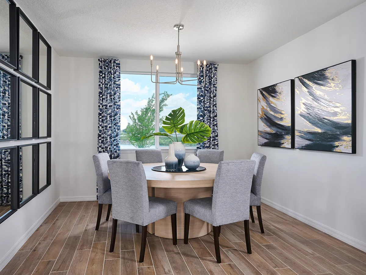Modern dining room with a round wooden table, six upholstered gray chairs, decorative vases with green leaves, abstract wall art, large window with patterned curtains, and wood-look tile flooring.