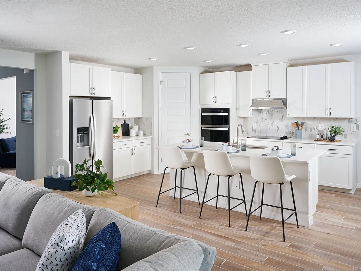Modern kitchen with white cabinets, stainless steel appliances, a marble backsplash, and a large island with four white barstools. Wooden floors and a gray sofa are visible in the open-concept space.