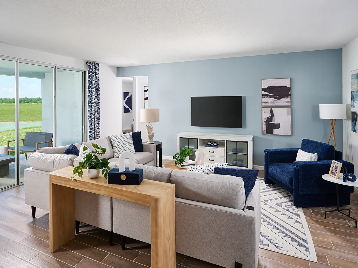 Modern living room with light gray sectional sofa, navy blue armchair, flat-screen TV on a white console, abstract wall art, floor lamp, wooden console table, indoor plants, and large glass doors overlooking greenery.