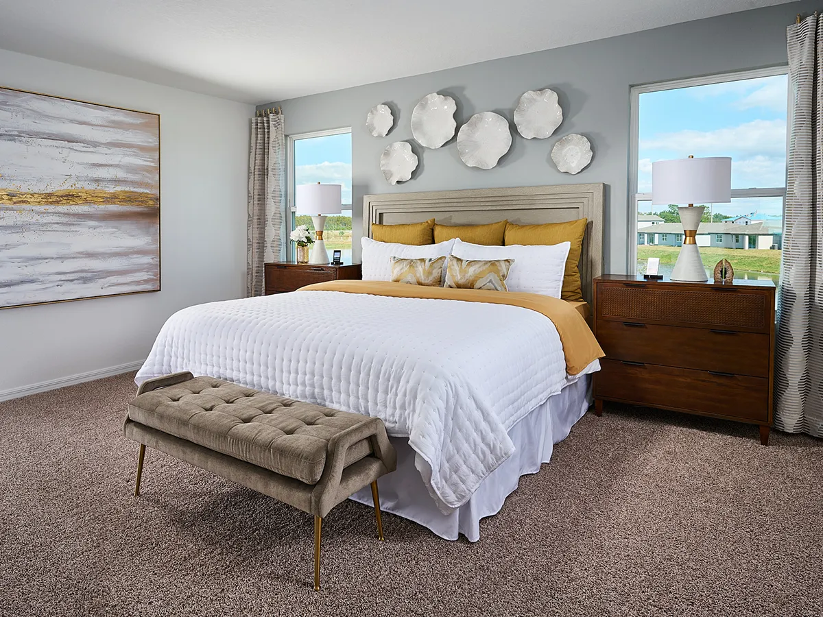 A modern bedroom with a large bed featuring white and gold bedding, a tufted bench at the foot, two wooden nightstands, large windows with curtains, abstract wall art, and decorative white wall sculptures above the headboard.