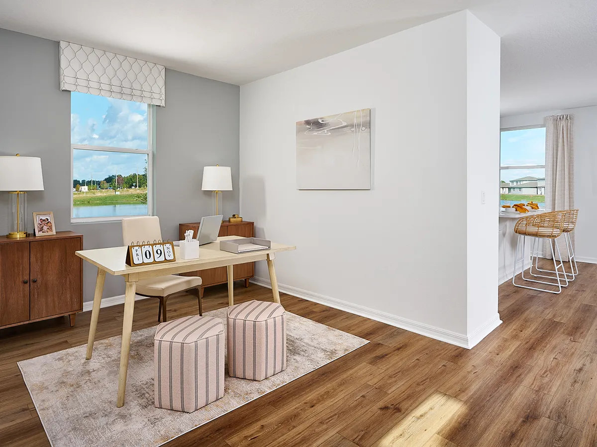 A modern home office features a wooden desk with two striped stools, a lamp, and framed photos. Theres a window, cabinet, and wall art. The room has wood flooring and a view of a bright, open kitchen area.