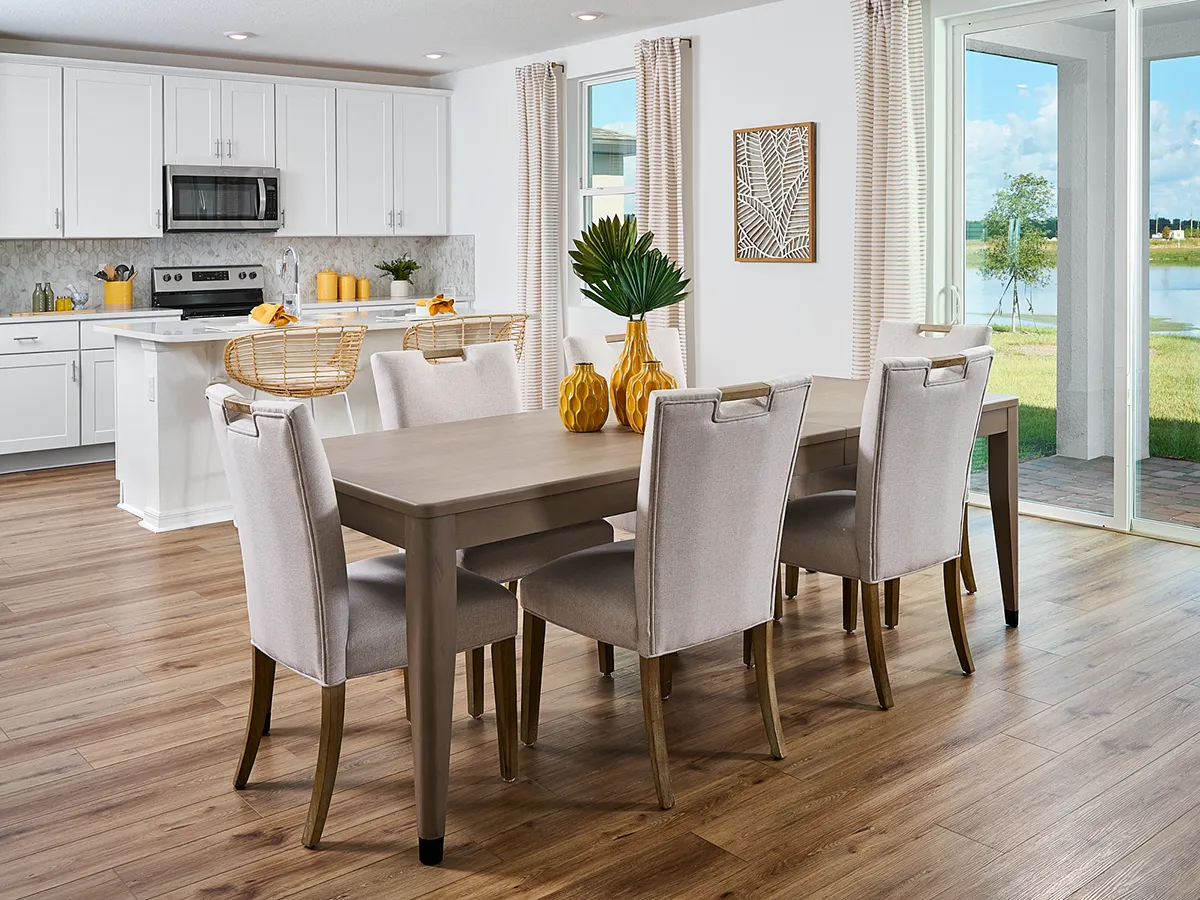 A modern dining room and kitchen with light wood floors, a long table with six upholstered chairs, white cabinets, stainless steel appliances, bar stools, and large windows showing a patio and a lake view.