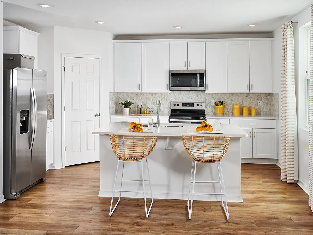 Modern kitchen with white cabinets, stainless steel appliances, a marble backsplash, and a central island with two wicker barstools. Yellow accents and wooden flooring add warmth to the bright space.