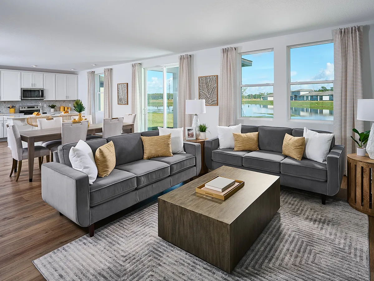 Modern living room with two gray sofas, a wooden coffee table, and neutral decor. Large windows let in natural light and reveal a lake view. Dining area and kitchen are visible in the background, featuring white cabinetry.
