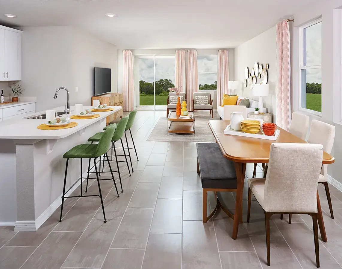 Modern open-concept kitchen and living area with a white island, green bar stools, wooden dining table, cream chairs, and a living room with large windows overlooking greenery. Neutral tones and bright natural light.
