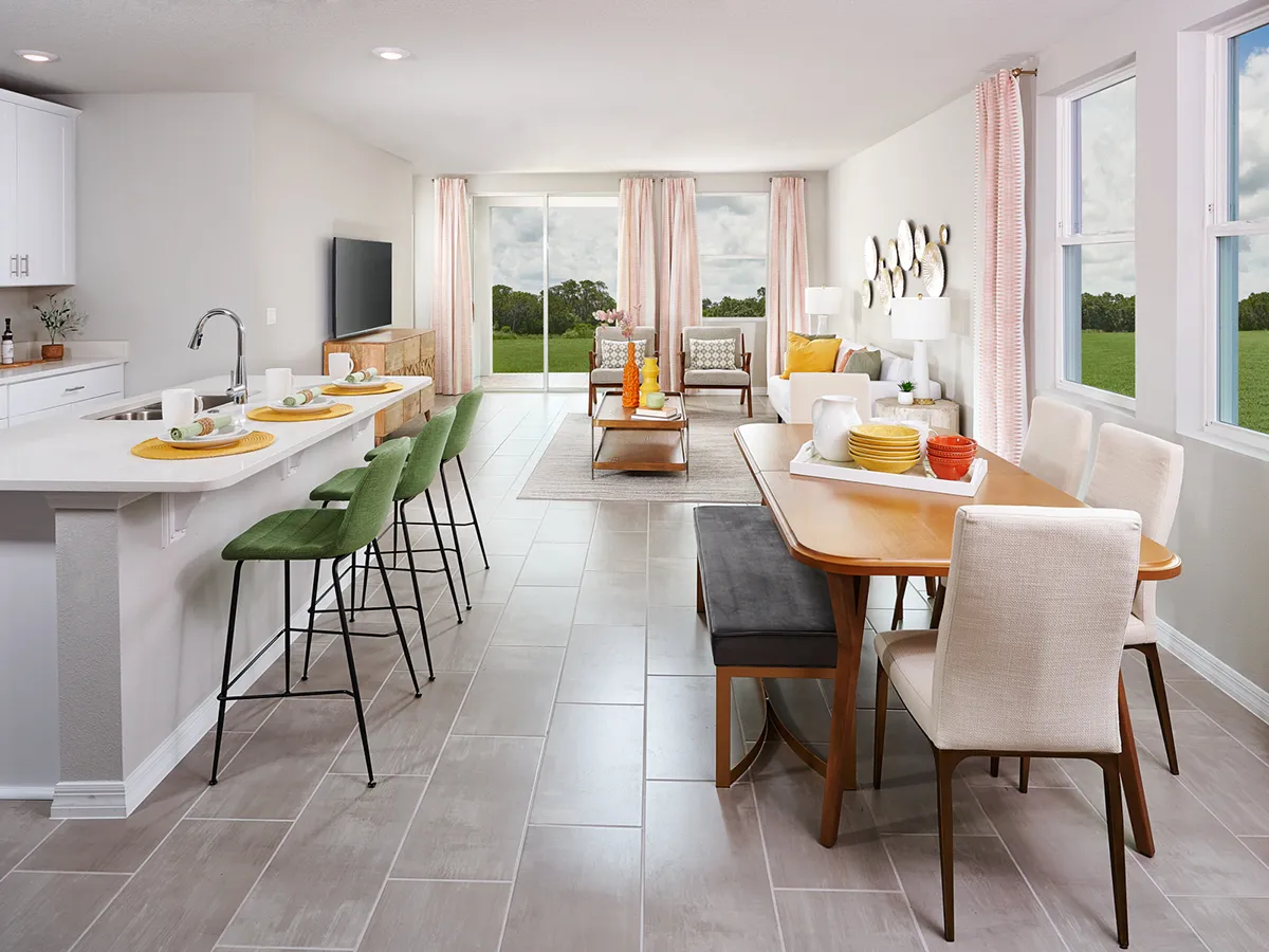 Modern open-concept kitchen and living area with a white island, green bar stools, wooden dining table, cream chairs, and a living room with large windows overlooking greenery. Neutral tones and bright natural light.