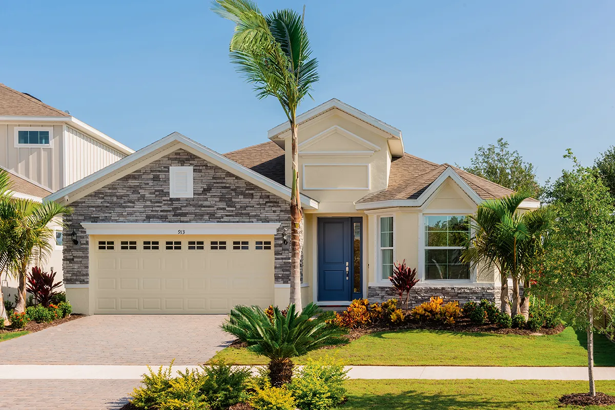Single-story suburban house with a double garage, stone and stucco exterior, blue front door, manicured lawn, palm trees, and various shrubs and plants in the front yard on a sunny day.