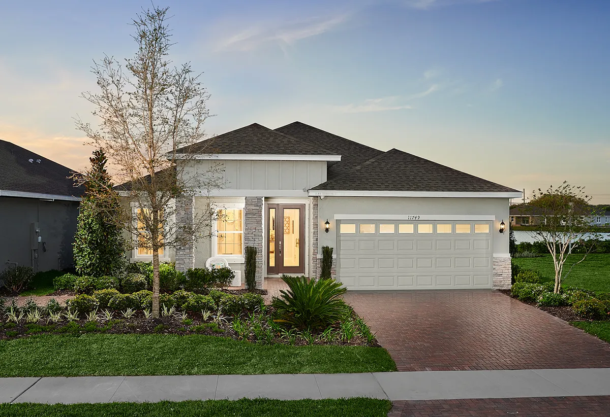 Modern single-story house with a brick driveway, double garage, and well-manicured front yard featuring green grass, shrubs, and a young tree, photographed at sunset with a clear sky.