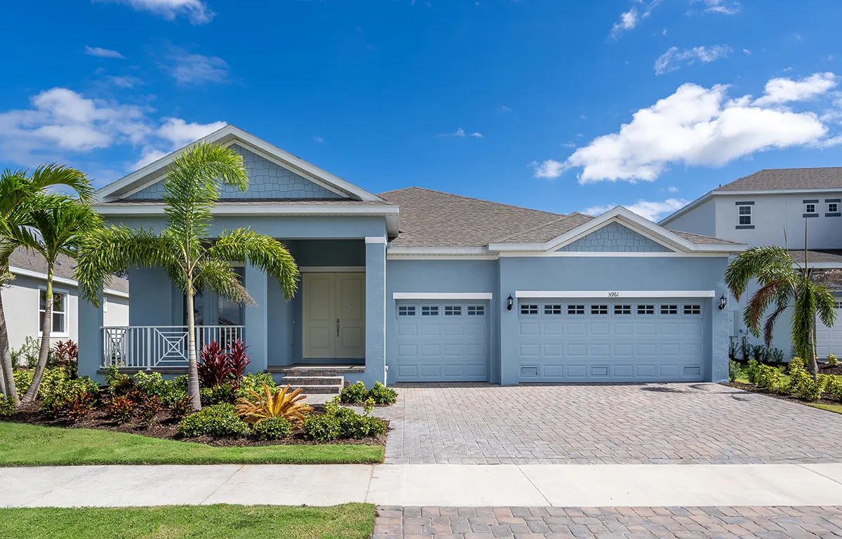 A modern light blue house with three garage doors, a front porch, palm trees, neatly trimmed lawn, and landscaped garden under a bright blue sky with scattered clouds.
